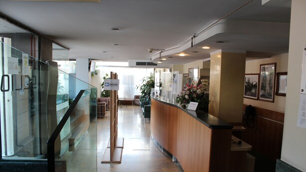 Uma área de recepção de hotel com balcão de madeira, guarda-corpos de vidro e escada para um andar superior.