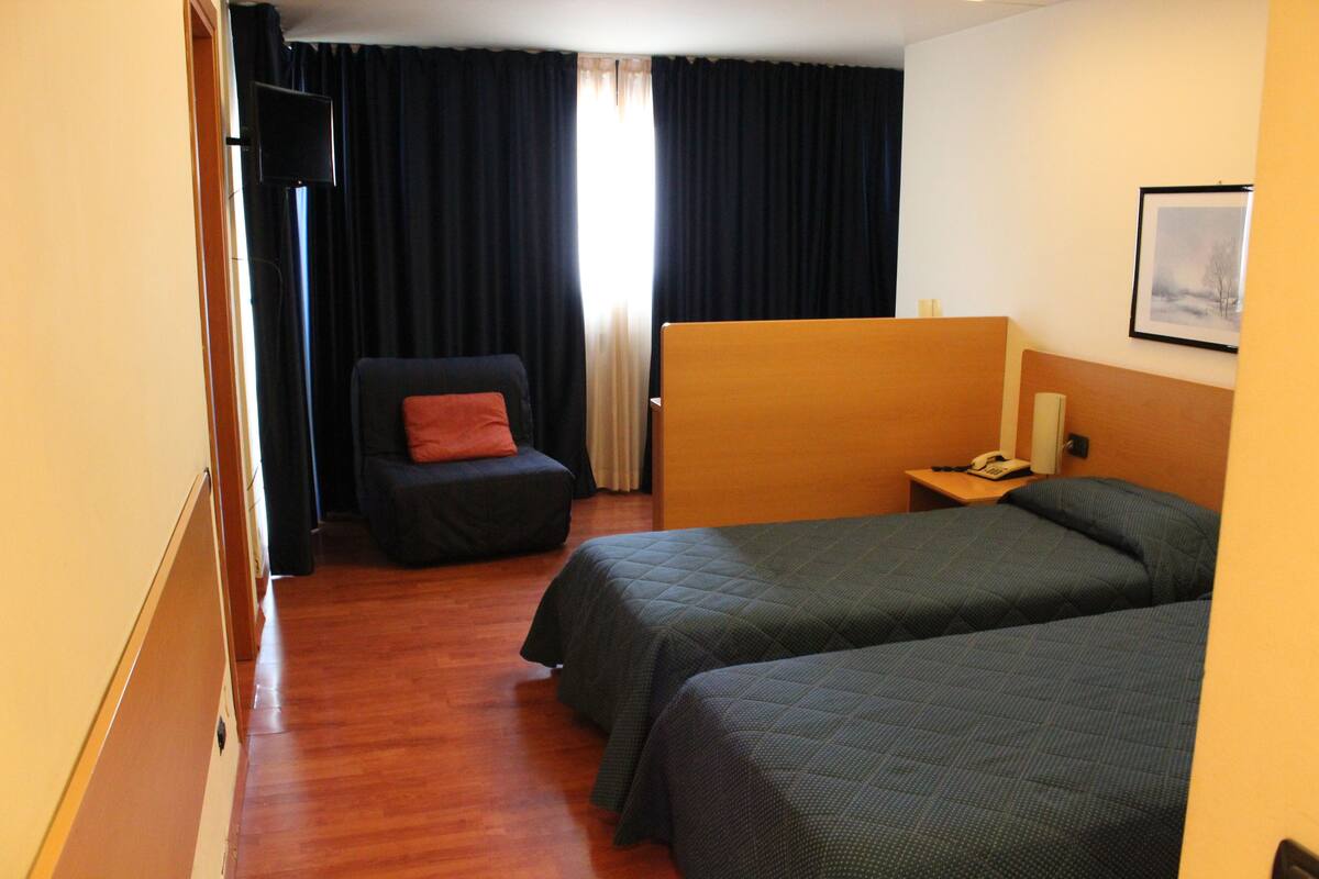 A hotel room with two beds, a chair, a desk with a phone, and a window with curtains.