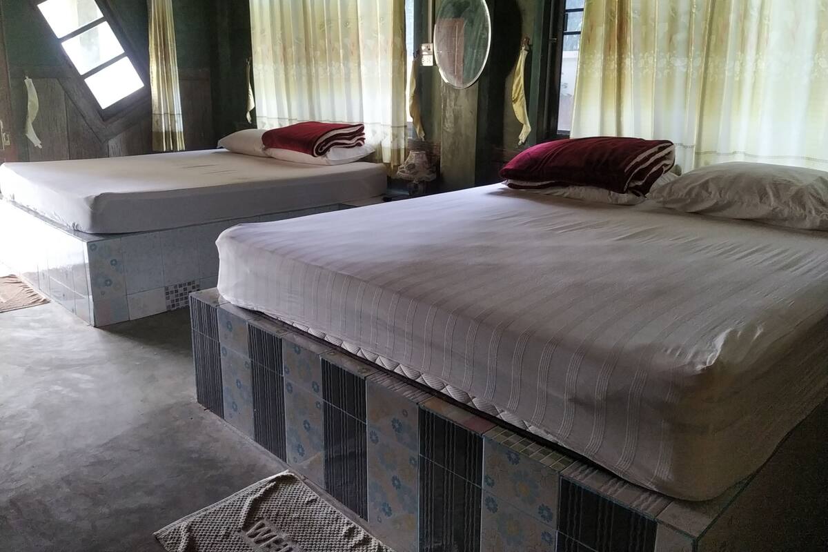 Duas camas de solteiro com roupa de cama branca e almofadas vermelhas, um espelho redondo pequeno e uma janela com cortinas finas.