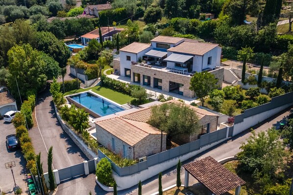 Una casa moderna de dos pisos con piscina, rodeada de exuberante vegetación y un camino de entrada pavimentado.