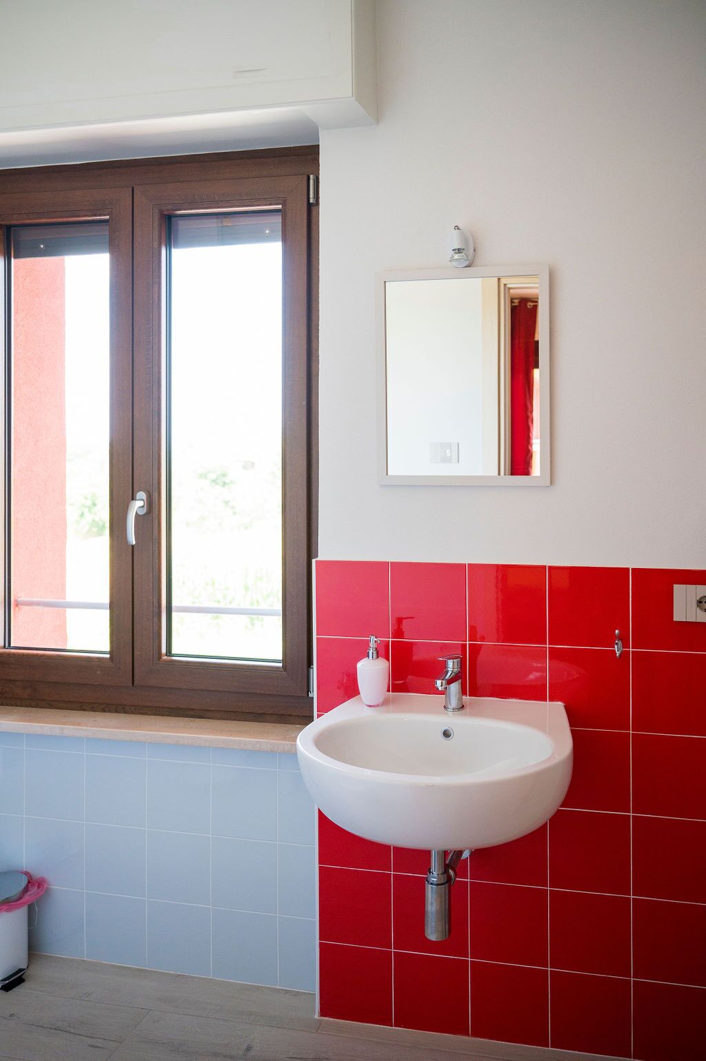 A bathroom with a white sink, red tiles, a window with a brown frame, and a mirror on the wall.