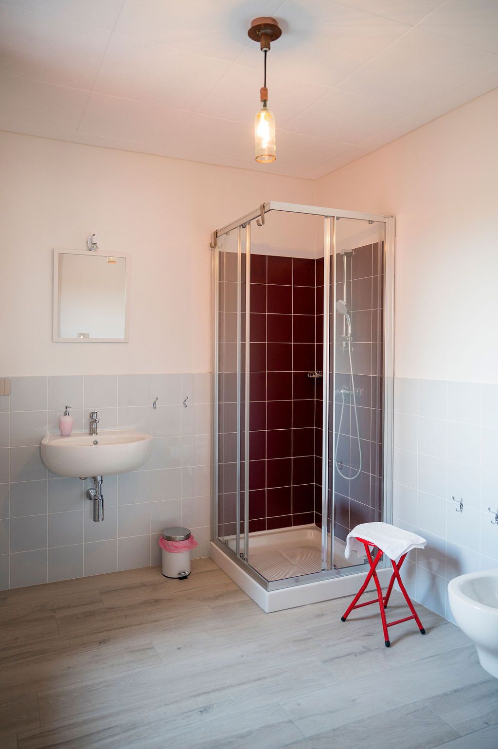 A modern bathroom with a glass-enclosed shower, a white sink, and a red stool.