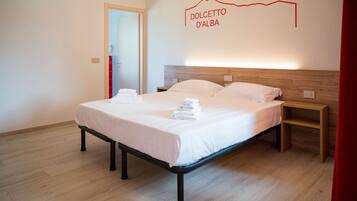 Un lit bien fait, avec du linge de lit et des serviettes blanches, une tête de lit en bois et une plaque fixée au mur portant l’inscription « DOLCETTO D’ALBA ».