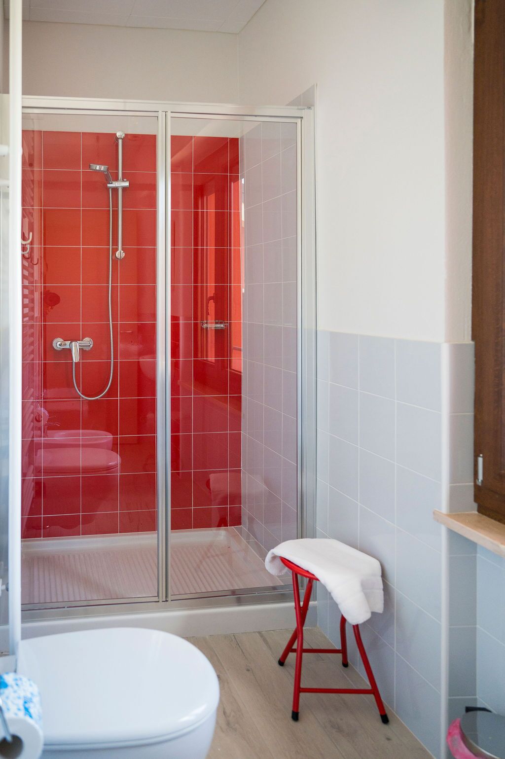 Een badkamer met een douchecabine van glas, een wit toilet en een rode kruk met een witte handdoek.