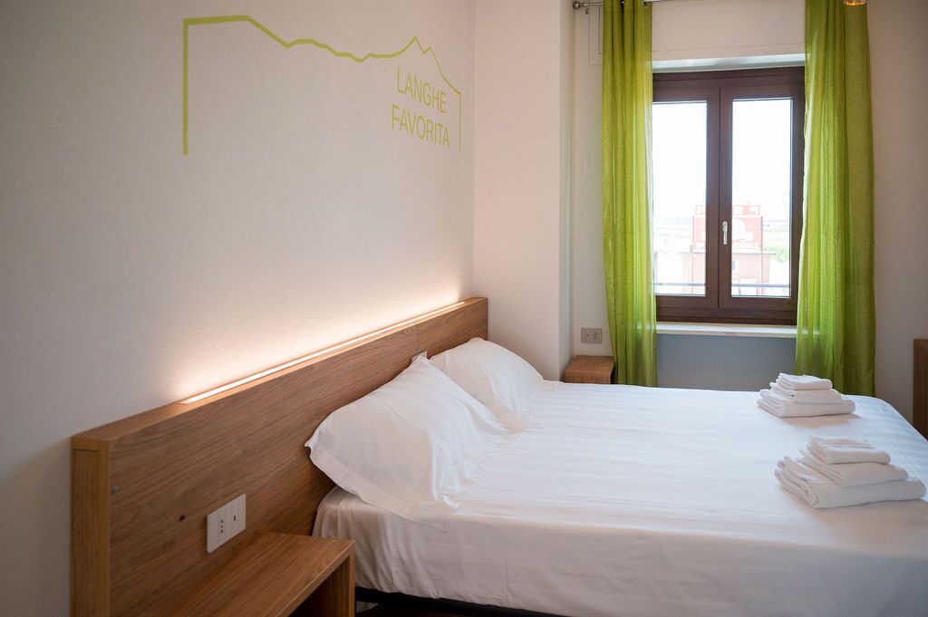 A hotel room with a neatly made bed, a headboard with a light switch, and a window with green curtains.