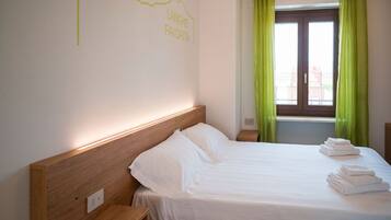 A hotel room with a neatly made bed, a headboard with a light switch, and a window with green curtains.