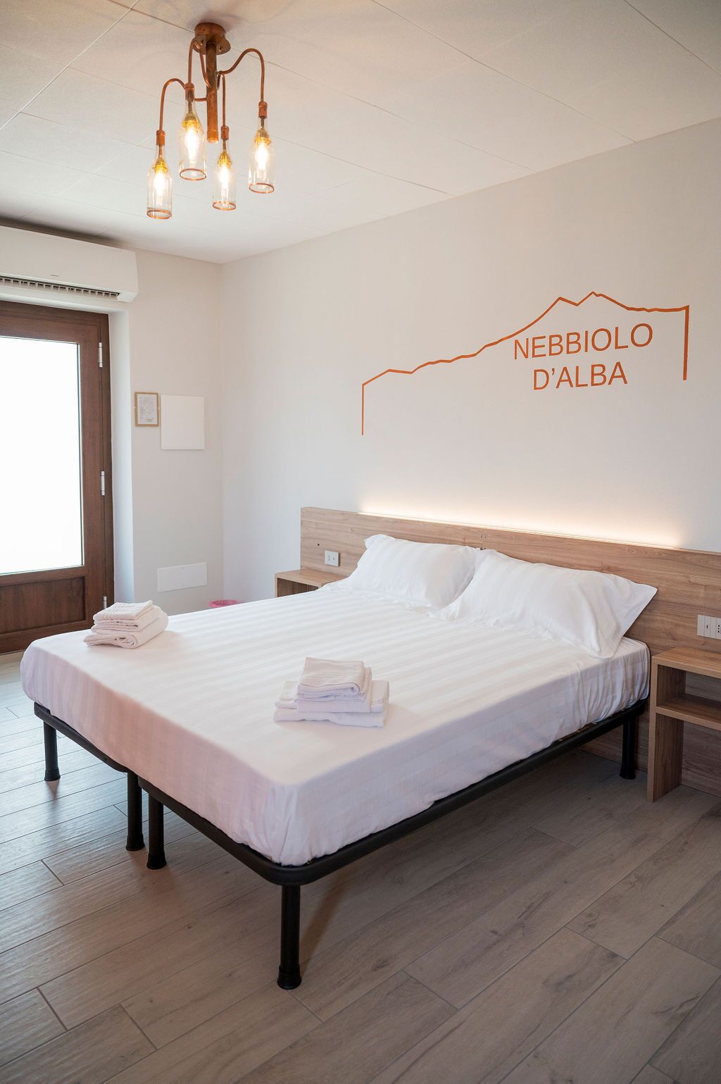 A neatly made bed with white linens and folded towels, a wooden headboard, and a wall-mounted light fixture.
