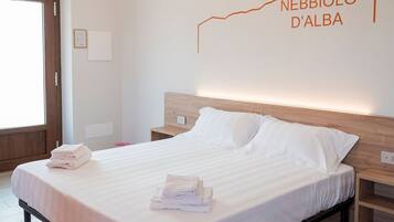 A neatly made bed with white linens and folded towels, a wooden headboard, and a wall-mounted light fixture.