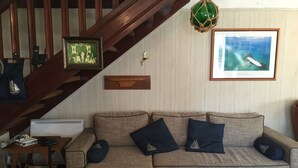 A living room with a wooden staircase, a sofa with cushions, a framed picture of a sailboat, and a framed picture of the Isles of Glénan.