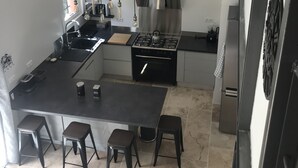 Een compacte keuken met een centraal kookeiland, een gasfornuis en een roestvrijstalen afzuigkap. Rond het eiland staan vier krukken en er is een klein spoelgedeelte.