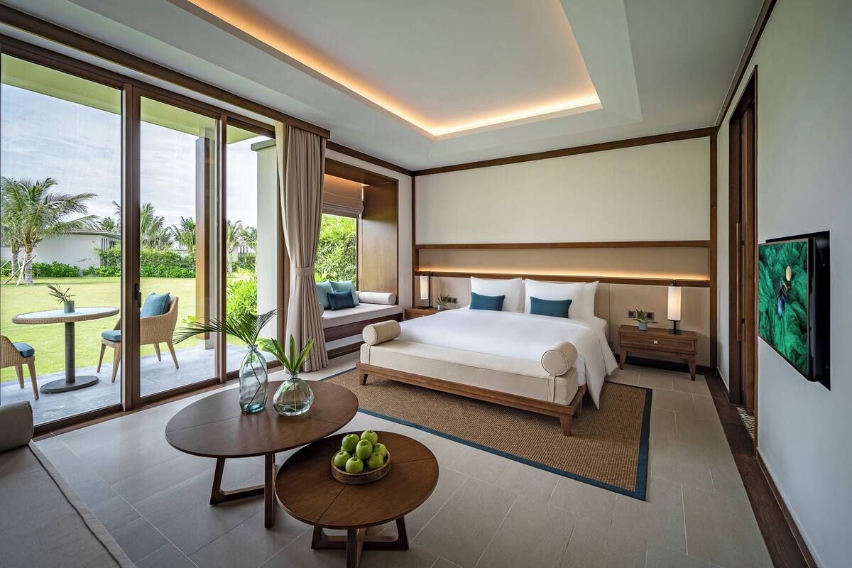 A modern hotel room with a large bed, a dining area with a table and chairs, and a view of the outdoors.