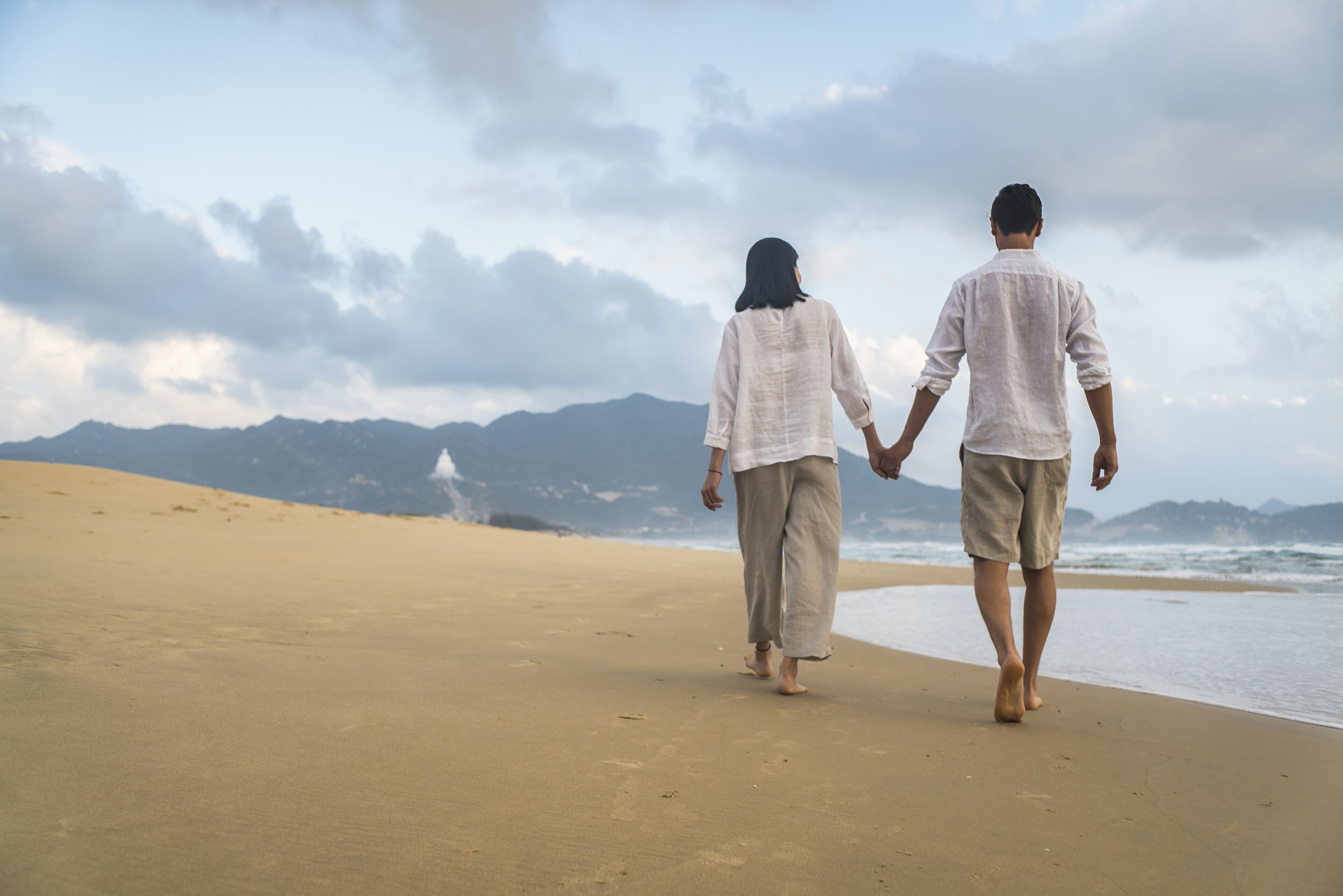A couple walking hand in hand on a sandy beach with mountains in the background.