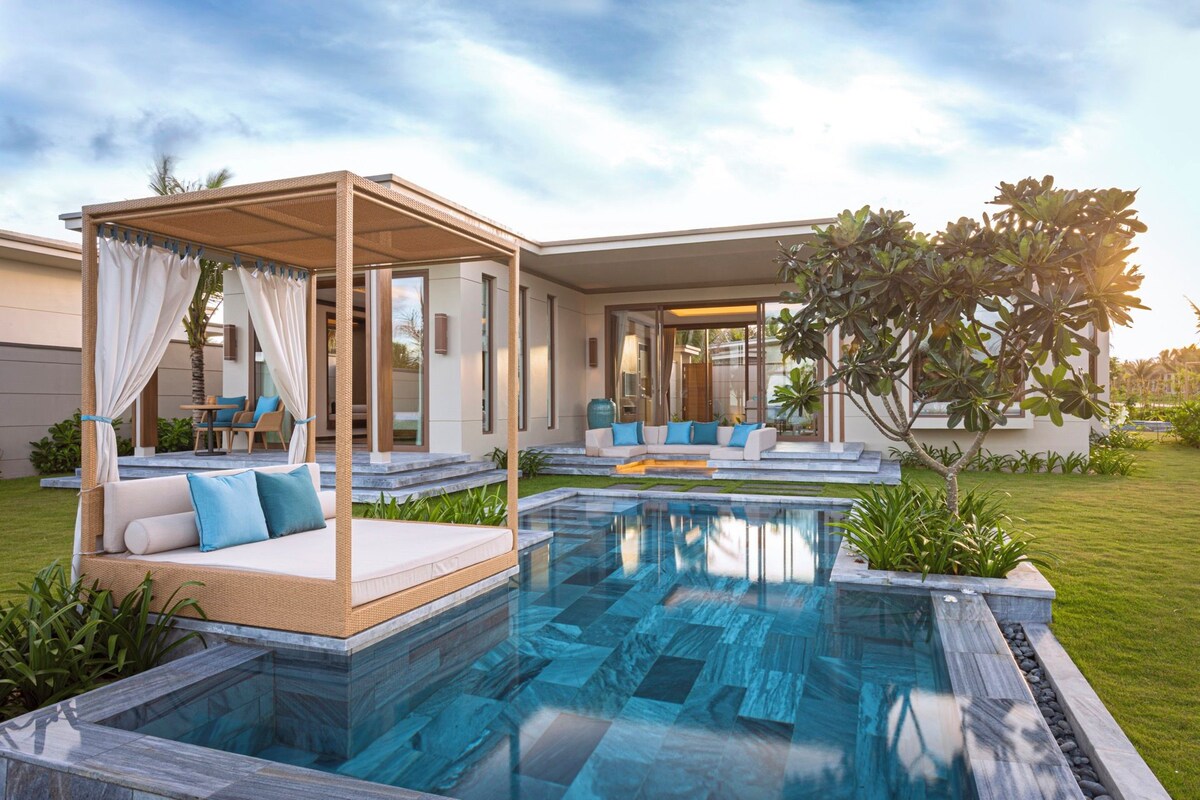Un espace piscine extérieur moderne doté d’un coin salon couvert, d’un salon avec des canapés et d’une pelouse bien entretenue.