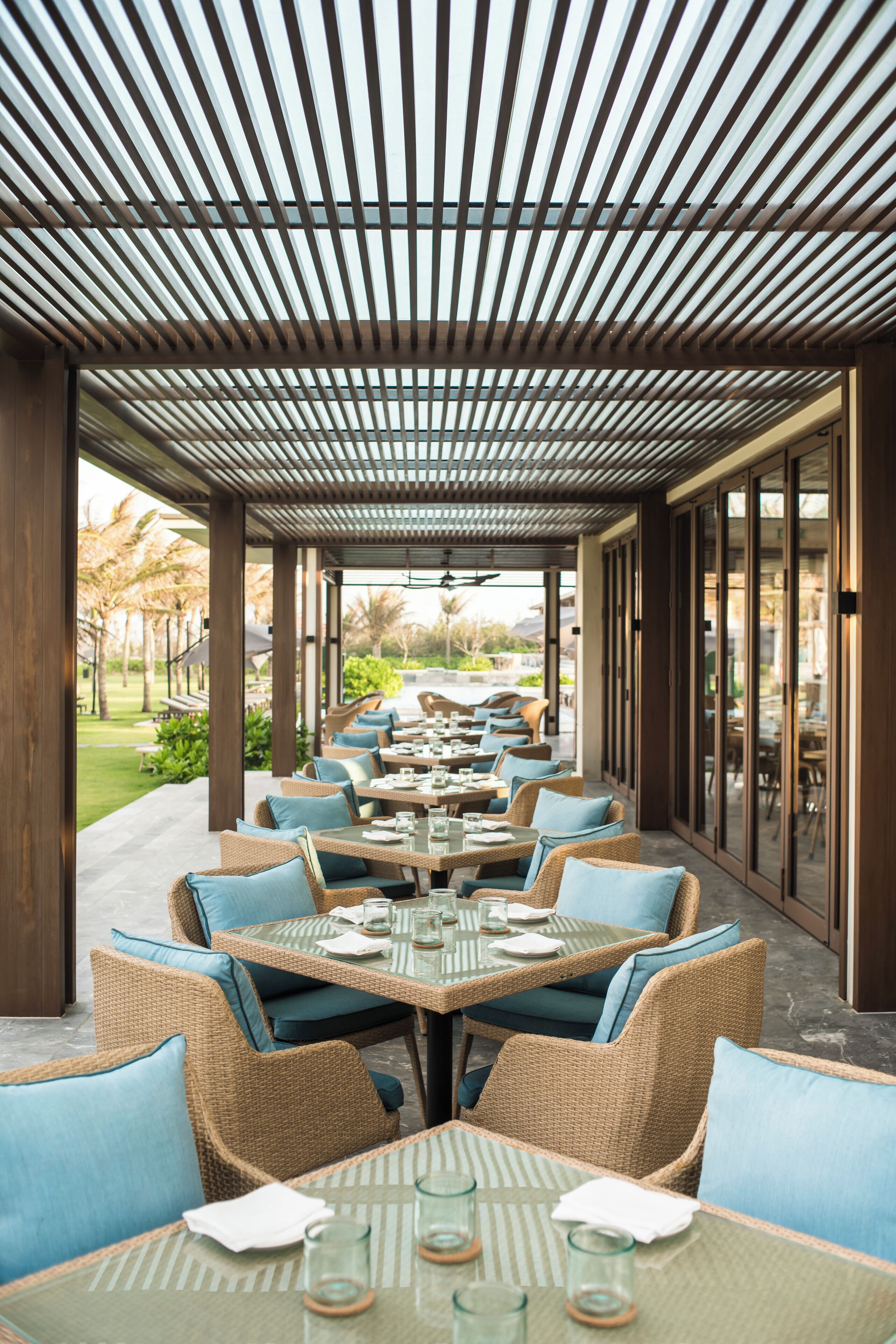 An outdoor dining area with wicker furniture and glass tables, featuring a covered roof and a view of the garden.