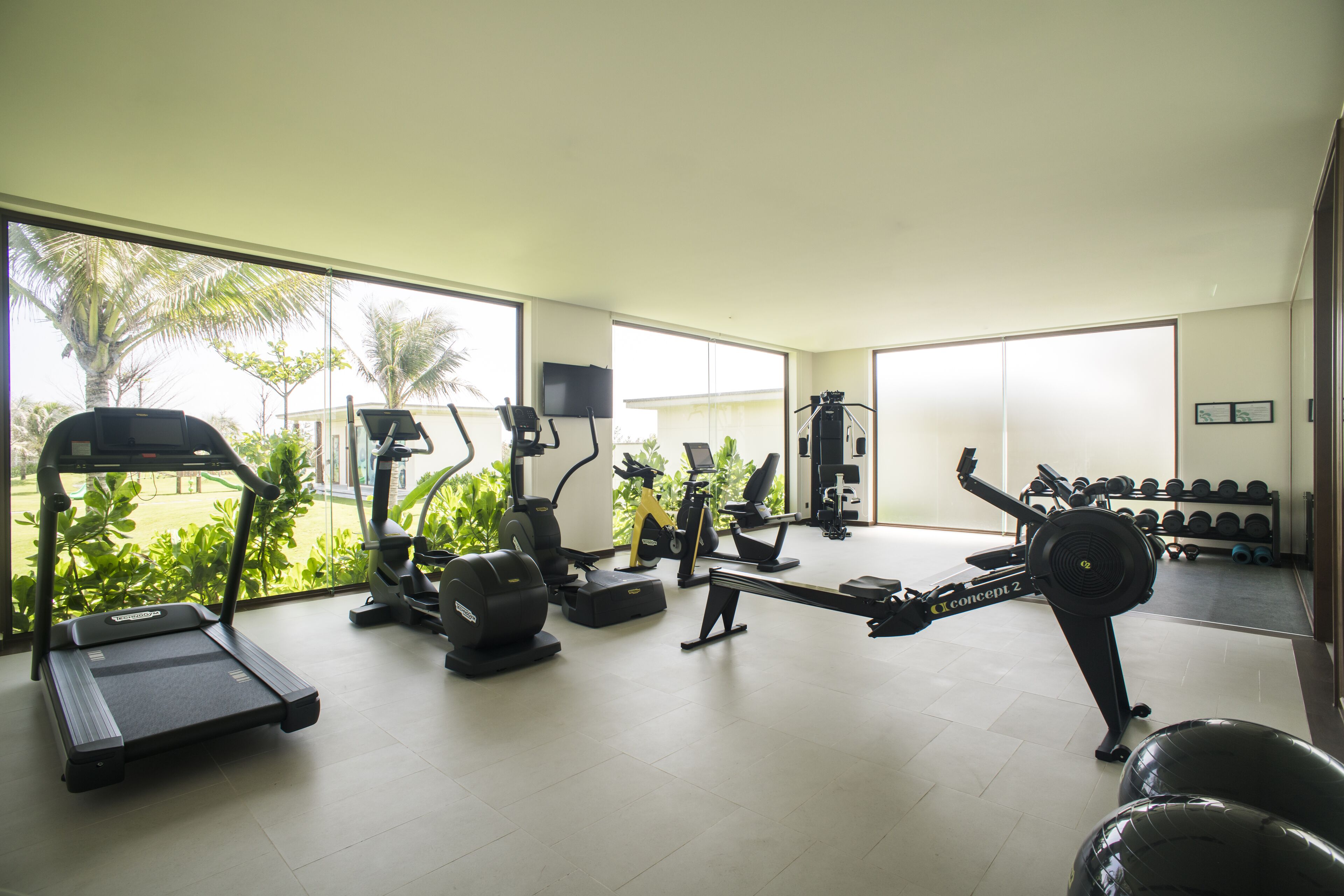 A gym with various exercise equipment, large windows, and a view of palm trees.