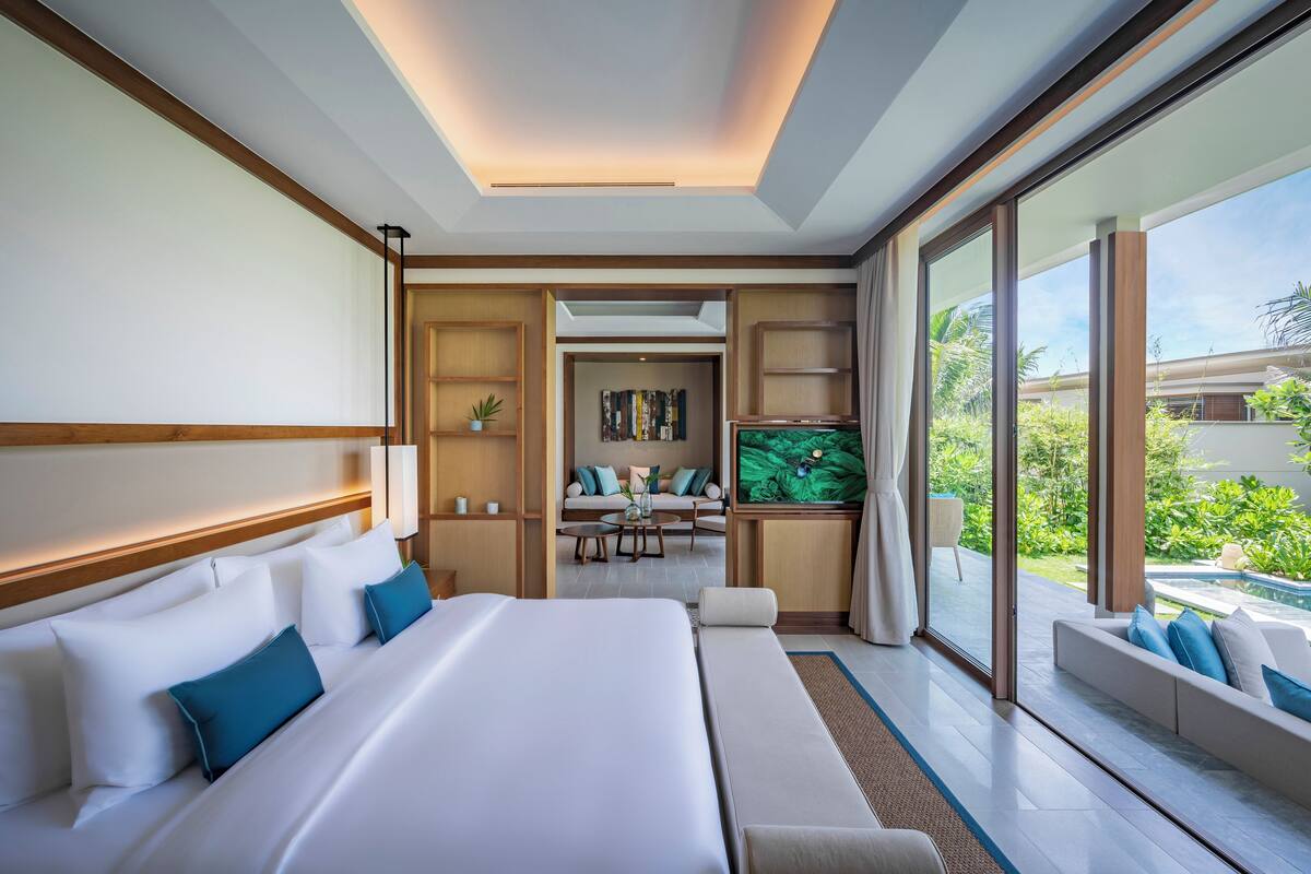 A modern hotel room with a large bed, a TV, and a view of a garden and pool.