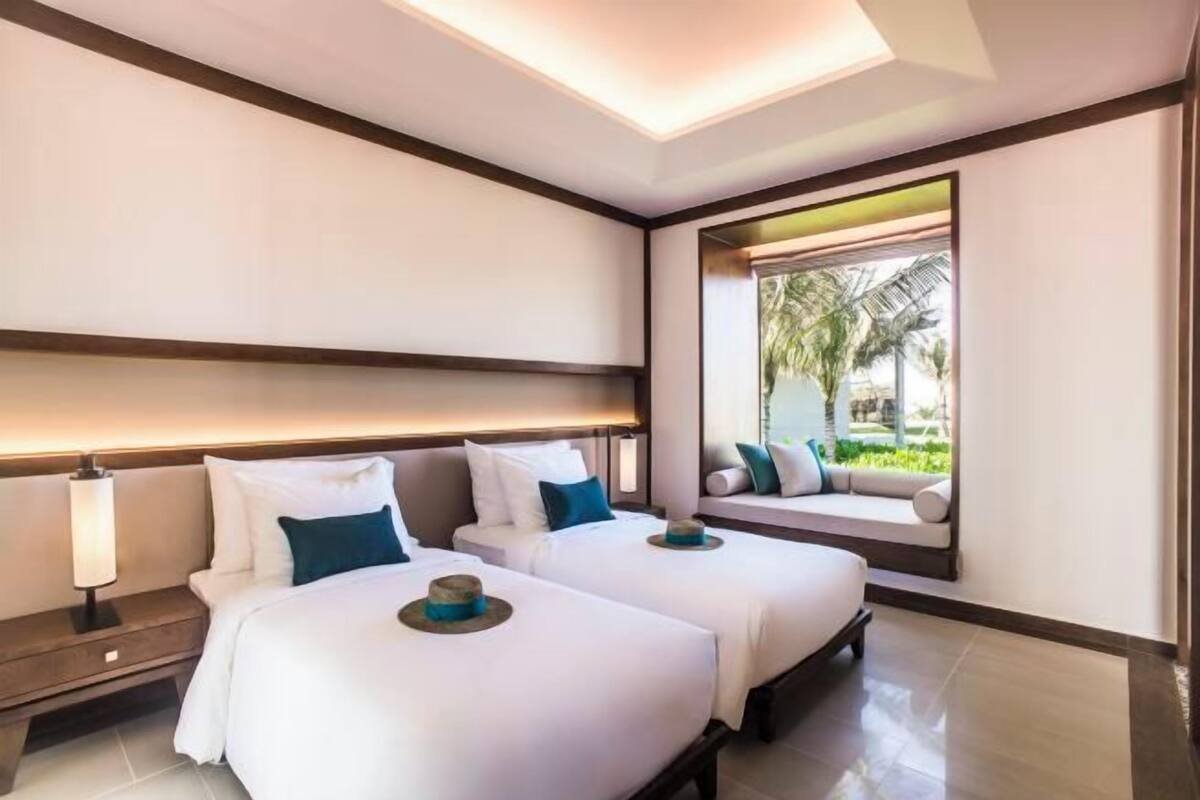 A hotel room with two beds, a nightstand, a lamp, and a window with a view of greenery.