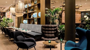 A modern lounge area with a sofa, chairs, and tables, featuring a wooden bookshelf and hanging pendant lights. - Motel One Bonn-Hauptbahnhof (Bonn)