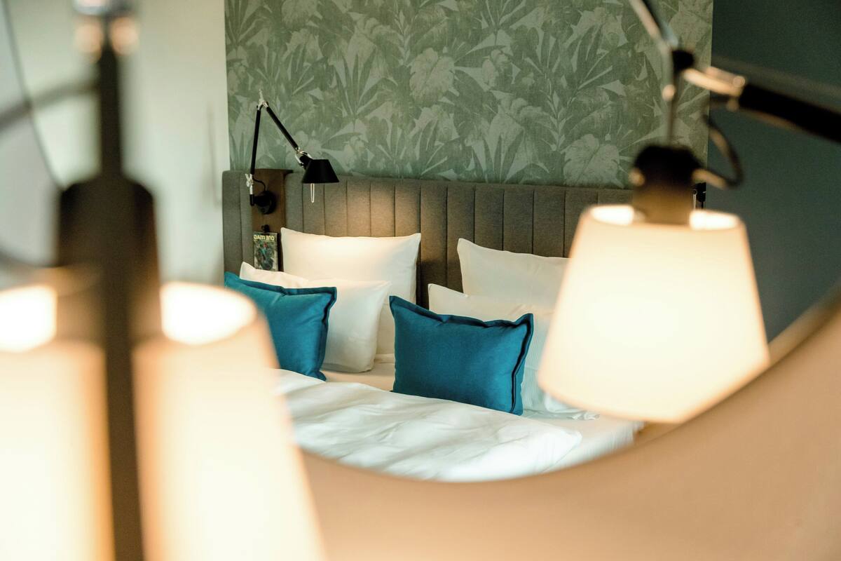 A neatly made bed with white and teal pillows, a grey headboard, and a green floral wallpaper.