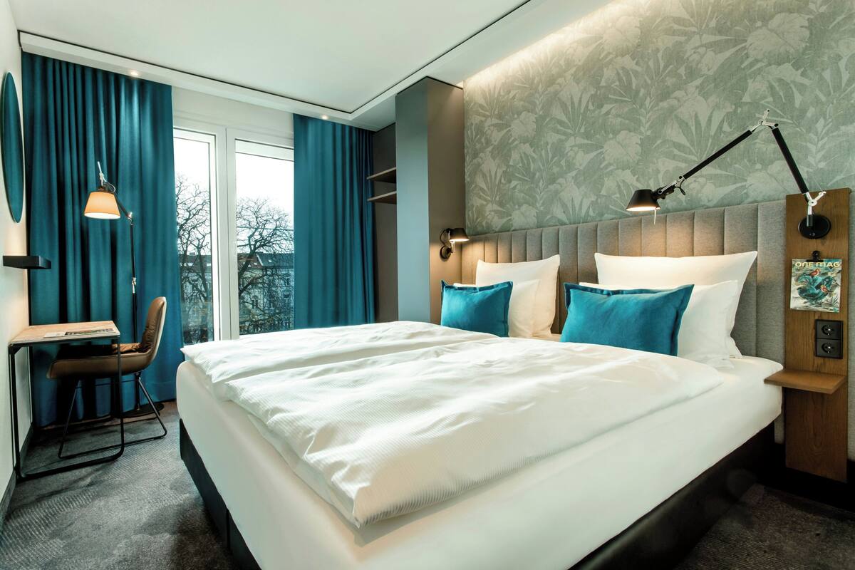 A hotel room with a large bed, a desk, and a chair. There is a window with curtains, a wall with a floral pattern, and a nightstand with a lamp.