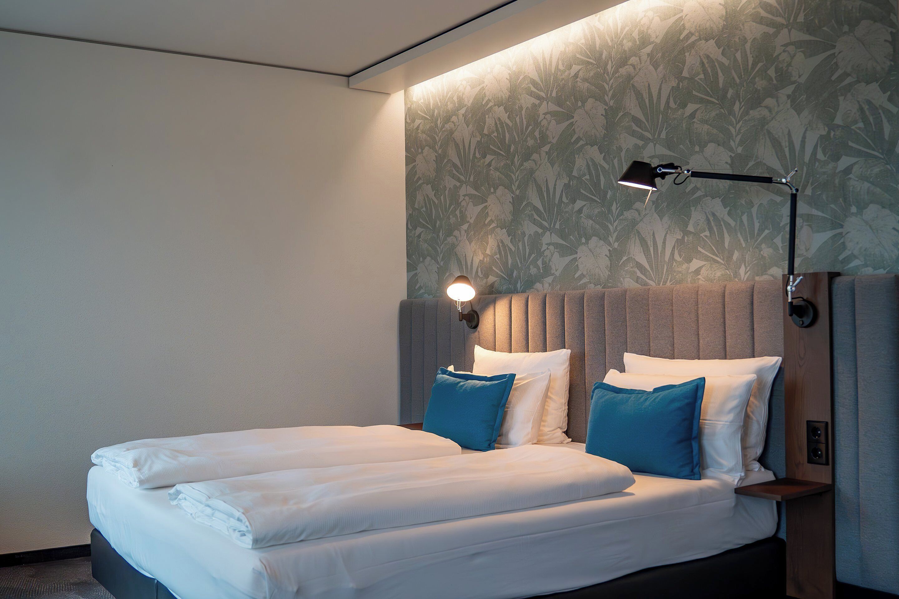 Una cama bien hecha con sábanas blancas y almohadas azules, un cabecero con una lámpara, y una pared con un diseño de hojas.