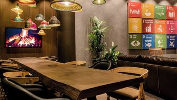 A modern dining area with a long wooden table, multiple chairs, hanging colorful lamps, a television displaying a fire, and a wall with the United Nations Sustainable Development Goals.