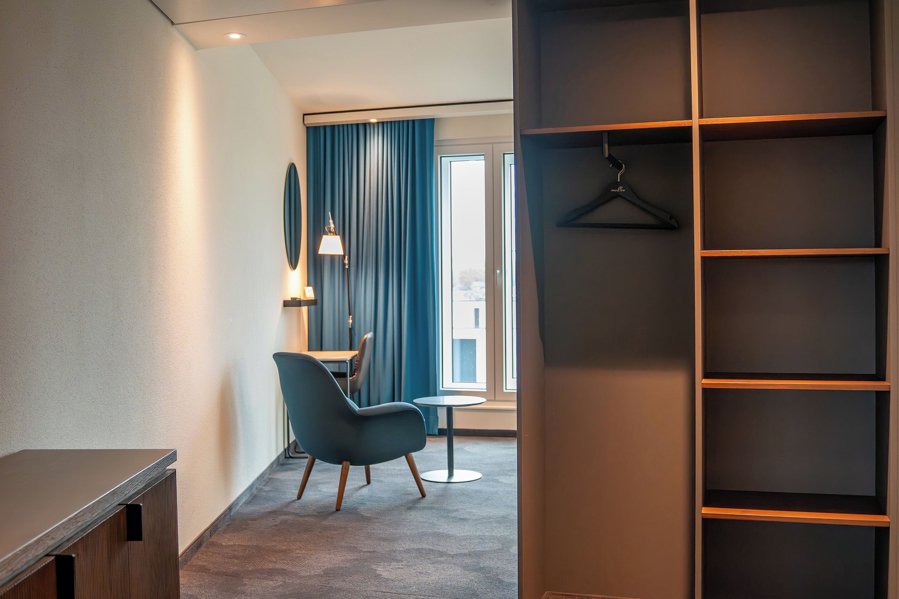 Habitación de hotel moderna con un armario grande, un escritorio, una silla y vistas a la ciudad a través de amplias ventanas.