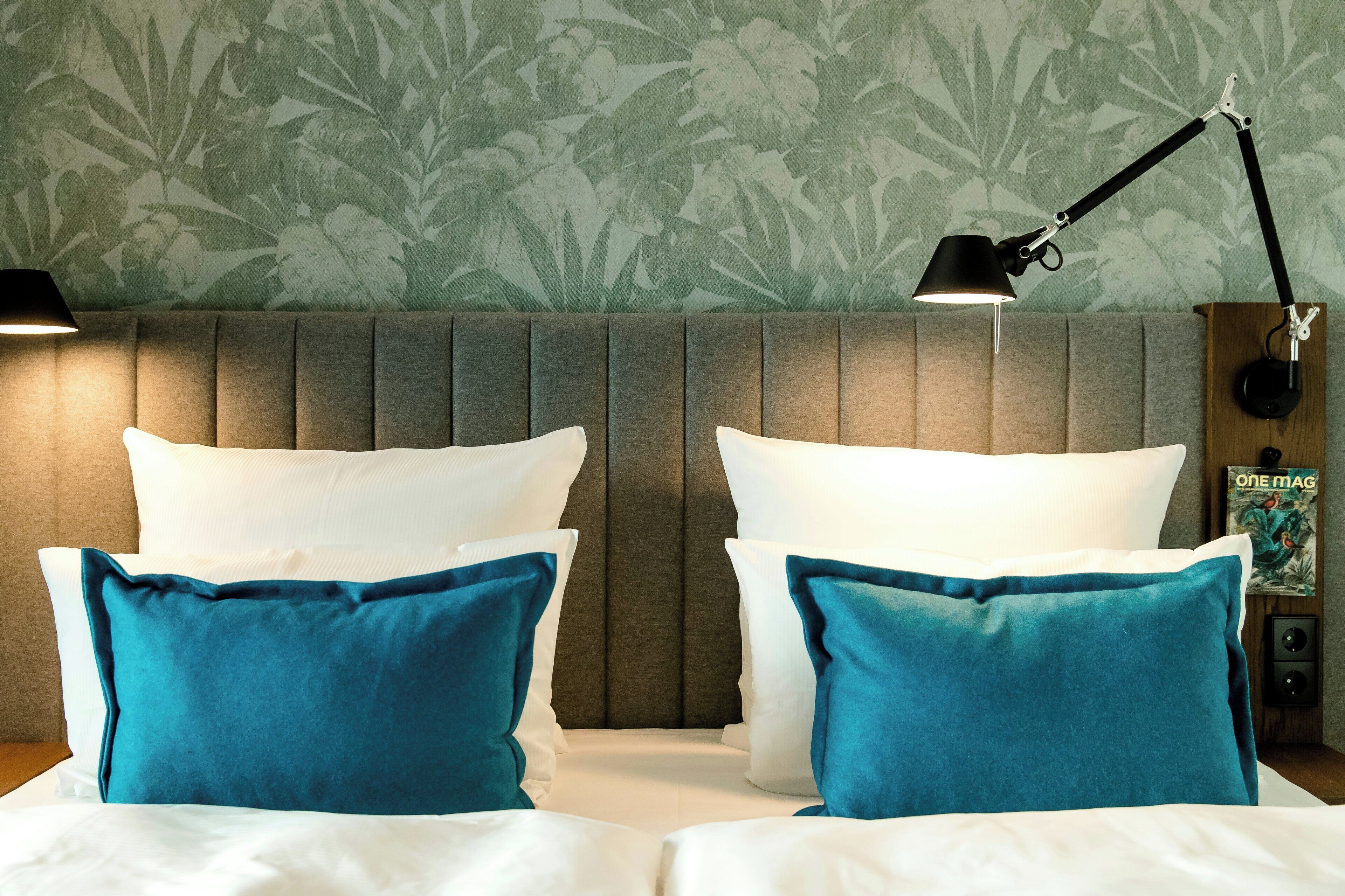 A hotel room with a bed, two pillows, a nightstand, a lamp, and a wall with a floral pattern.