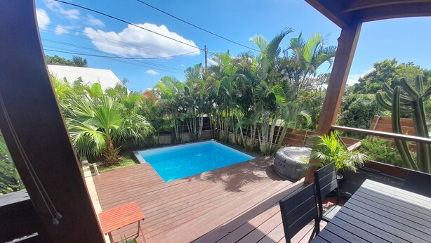A wooden deck with a pool, surrounded by tropical plants and a view of a house in the background. - KAZ VILLELE 4 STARS Pool / Jacuzzi (Saint Gilles les hauts)