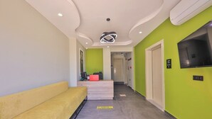 A reception area with a yellow sofa, a reception desk, a modern light fixture, a television, and a green wall. - Zeytin Hotel (Kas)