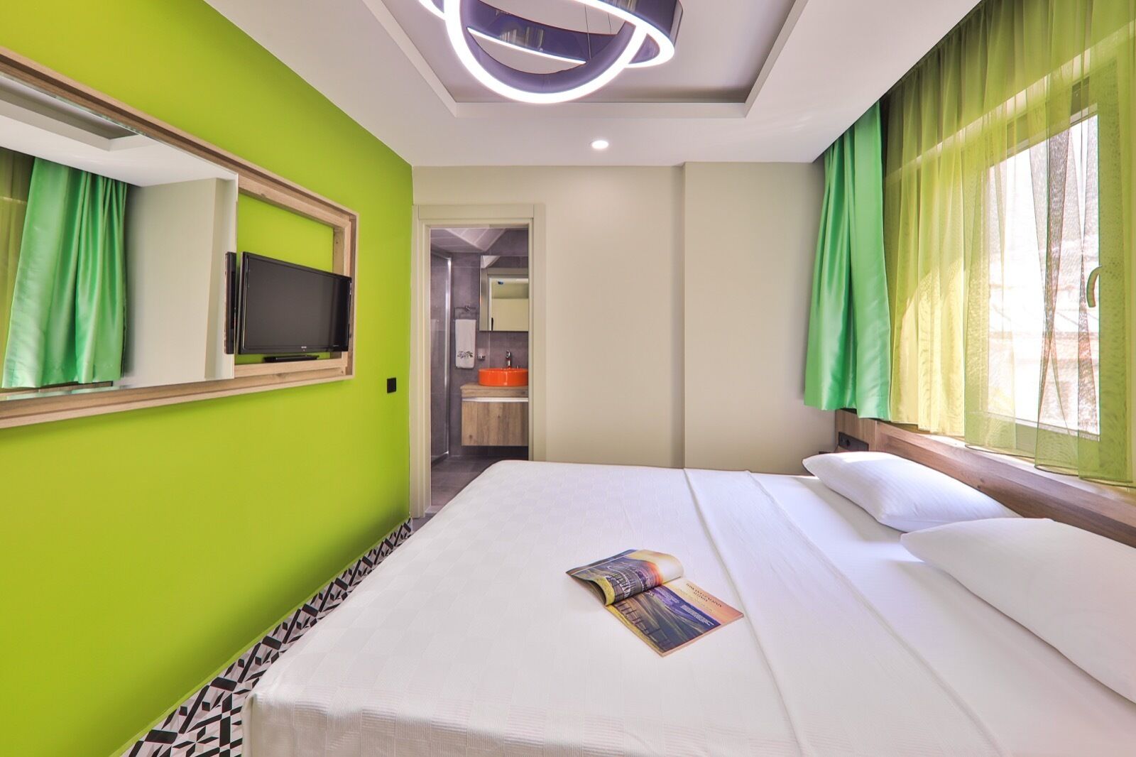 A hotel room with a large bed, a television, a bathroom with an orange sink, and green curtains.