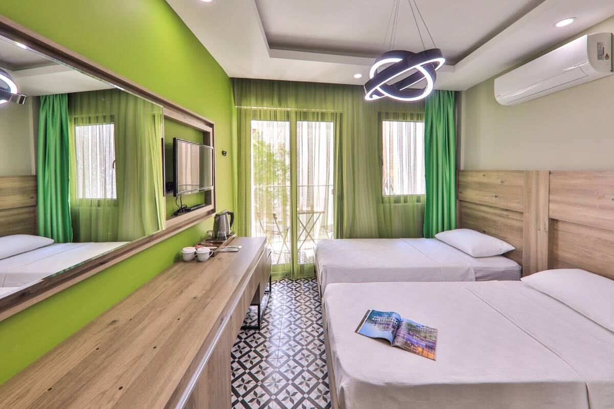 Habitación de hotel con dos camas, un escritorio de madera, un espejo, televisión y un ventanal con cortinas verdes.