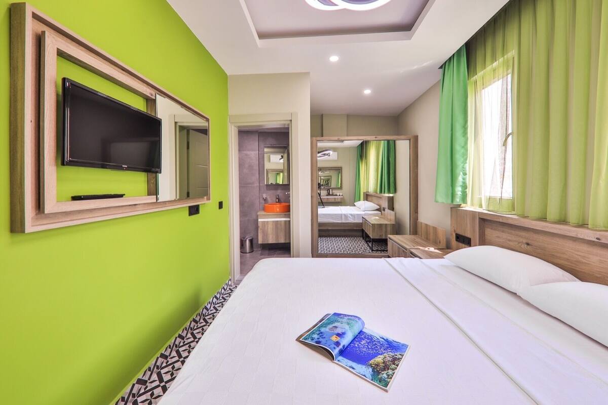 Una habitación de hotel con una cama grande, un televisor montado en la pared, un baño con lavabo y espejo, y cortinas verdes.