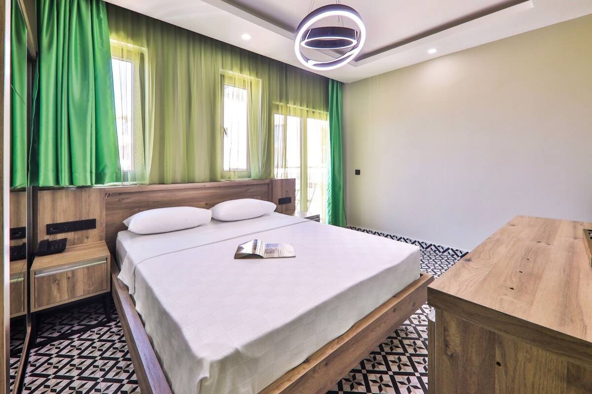 Una habitación de hotel moderna con una cama de madera, cortinas verdes, una mesita de noche pequeña y un suelo estampado.