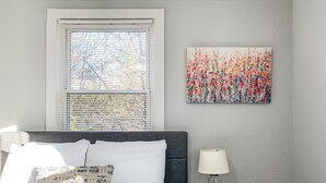 Une chambre à coucher avec un lit, une fenêtre avec des stores, une lampe fixée au mur et un tableau représentant des fleurs.