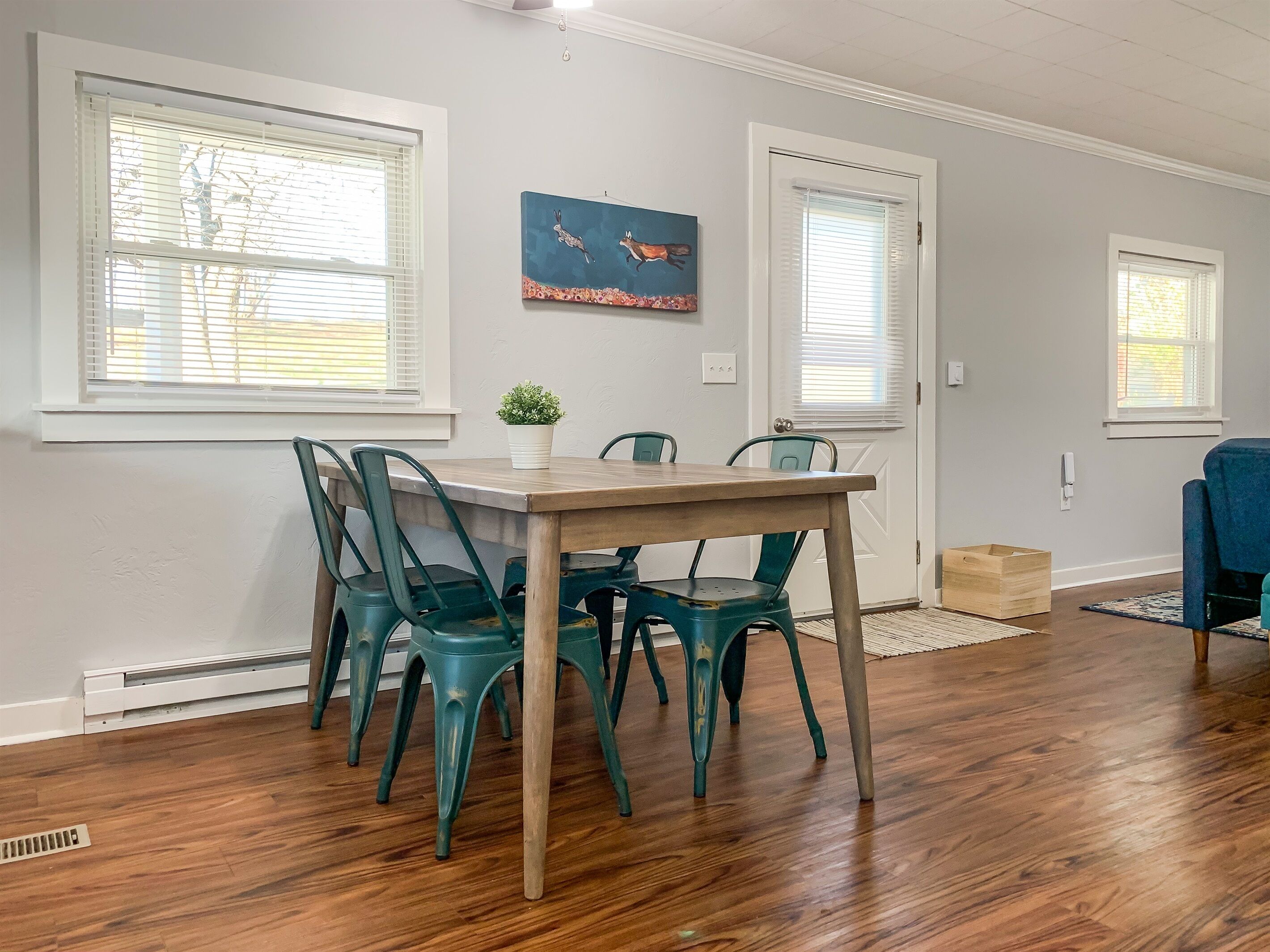 A dining area with a wooden table and teal chairs, a potted plant, and a painting on the wall.