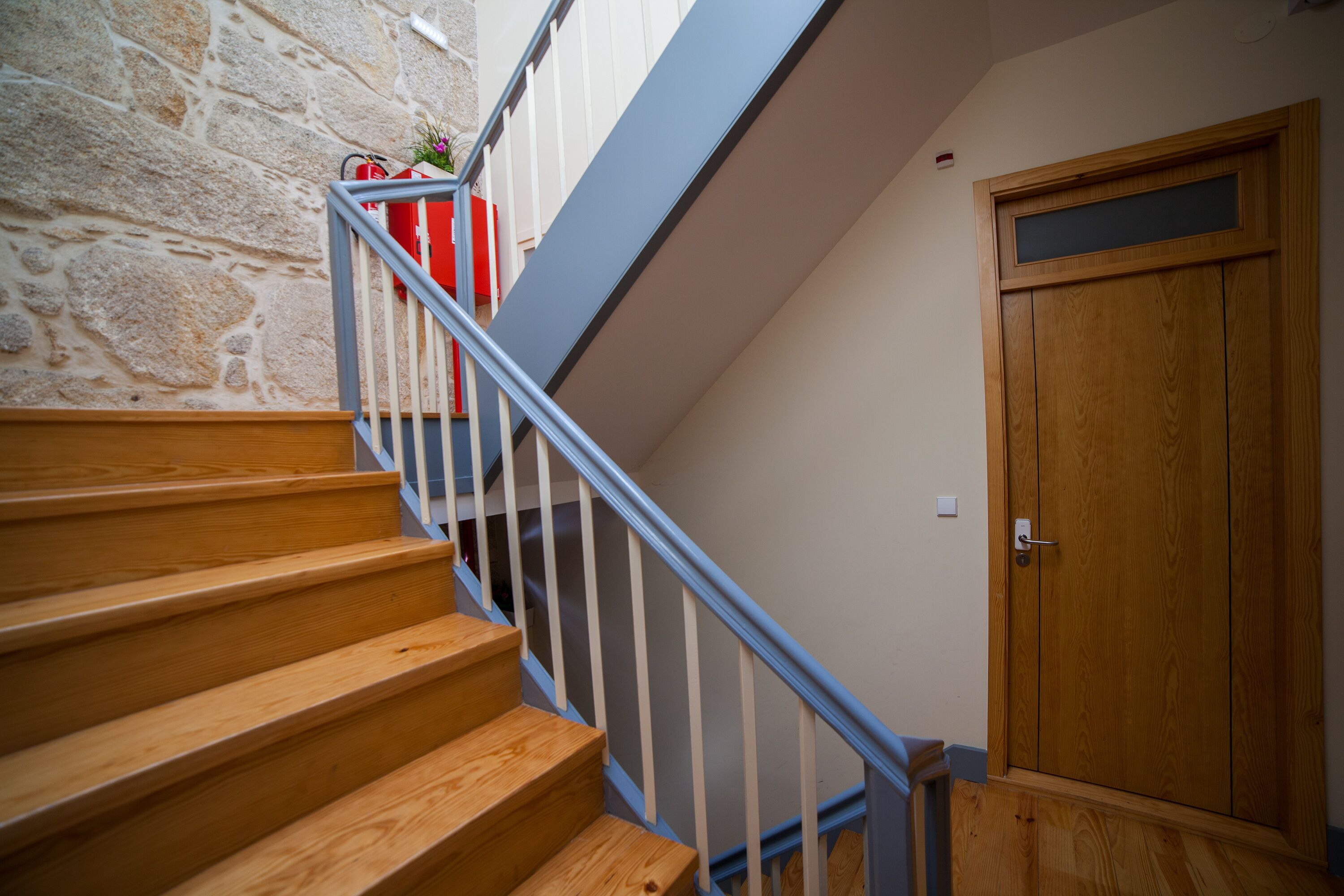Una escalera de madera con barandilla azul que conduce a una habitación con puerta de madera y pared de piedra.
