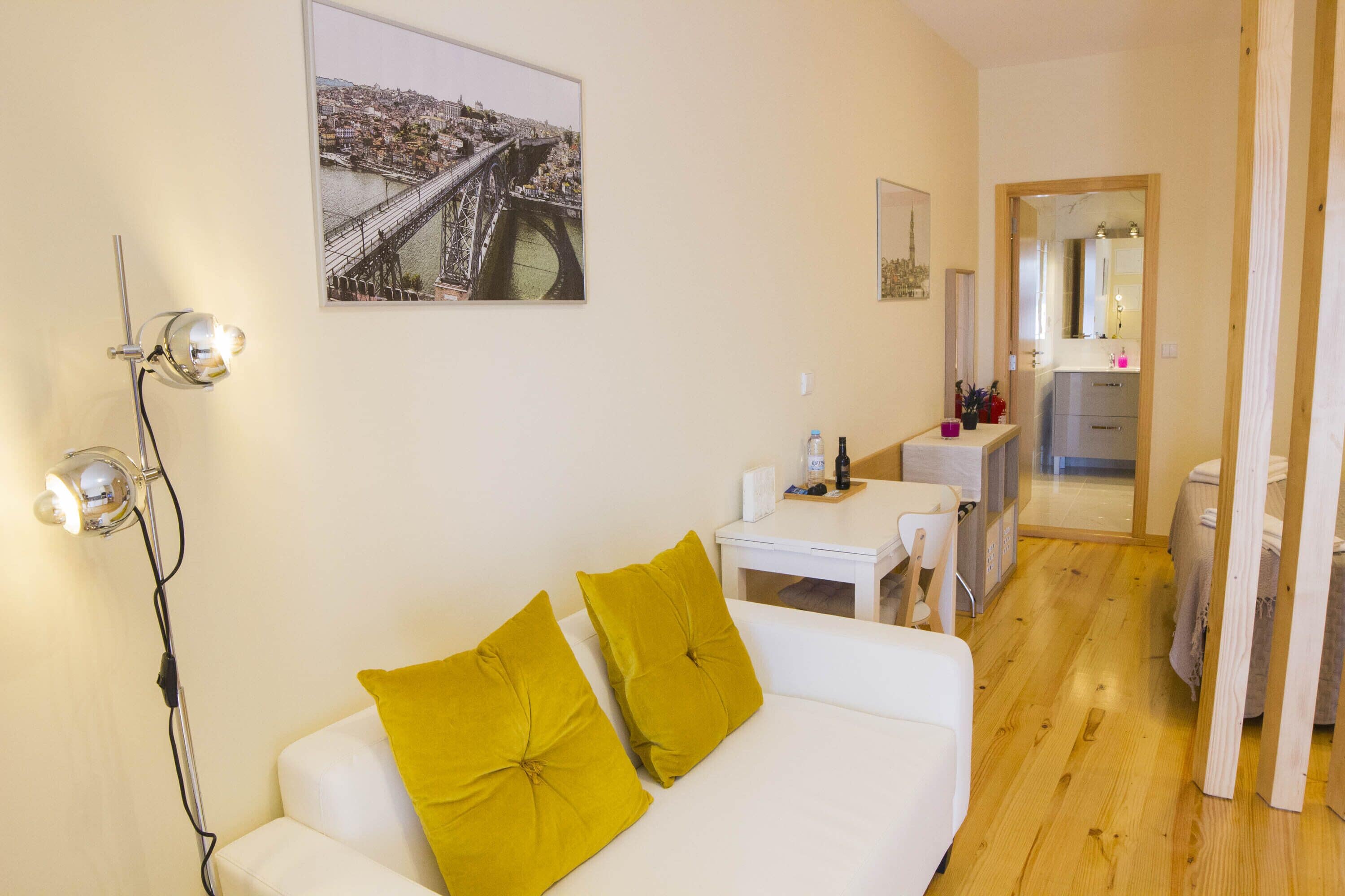 Un salon moderne avec un canapé blanc, deux coussins de couleur jaune moutarde, une lampe sur pied et un tableau représentant un paysage urbain, encadré et accroché au mur.