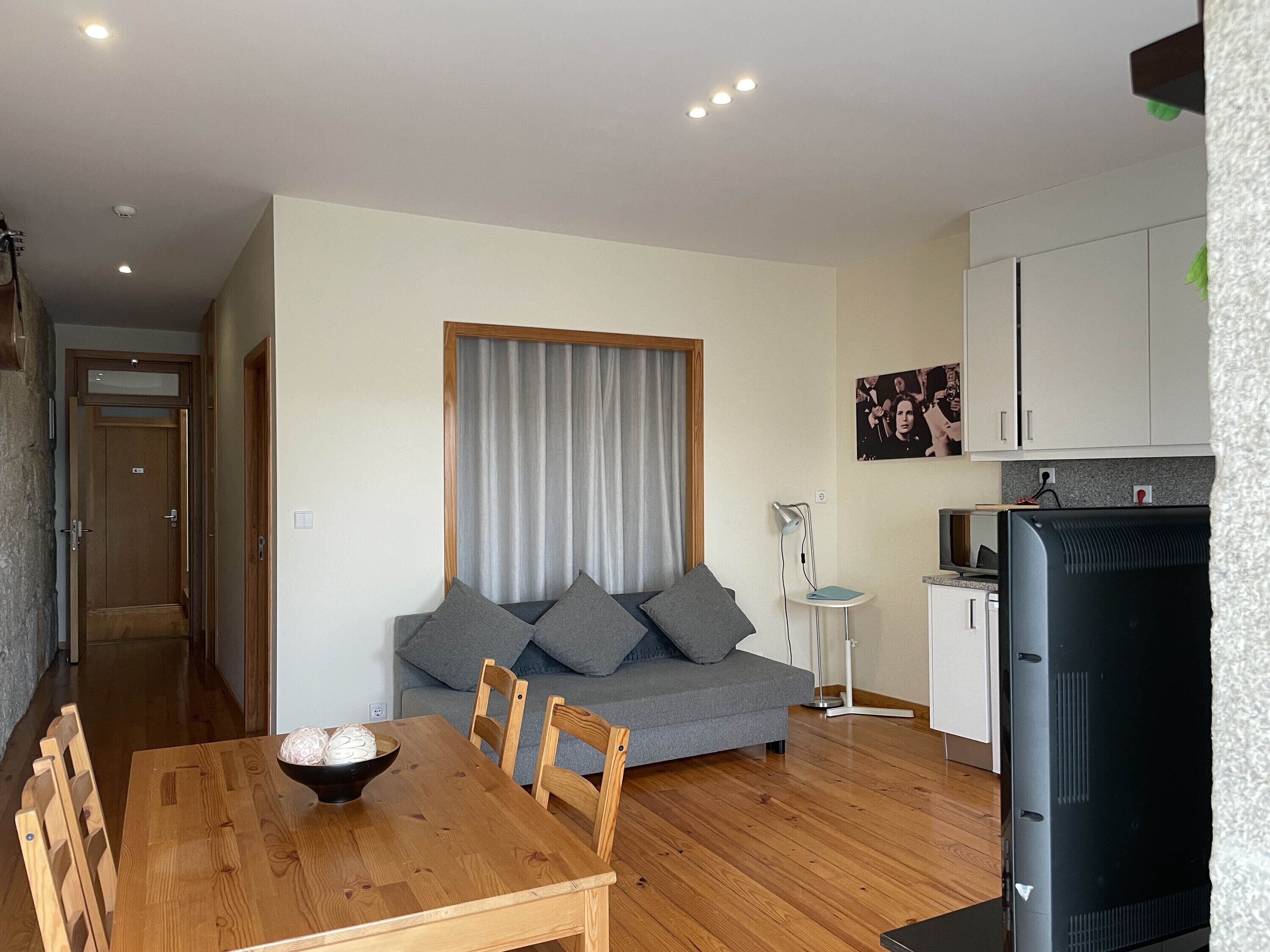A modern living room with a grey sofa, wooden dining table, and chairs. There is a small kitchen area with a fridge and a lamp.