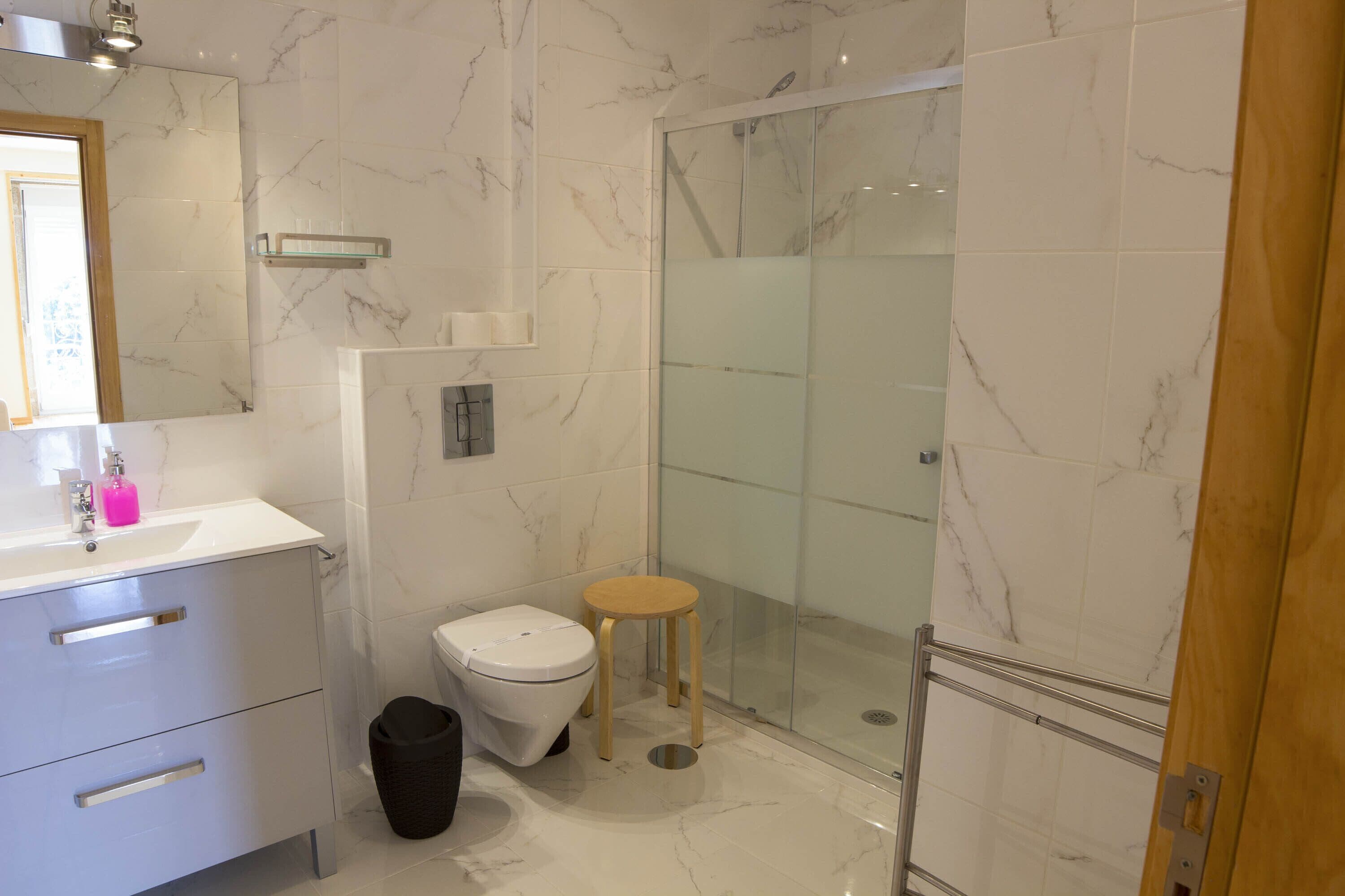 A modern bathroom with a marble floor, a glass shower enclosure, a wooden stool, and a white toilet.