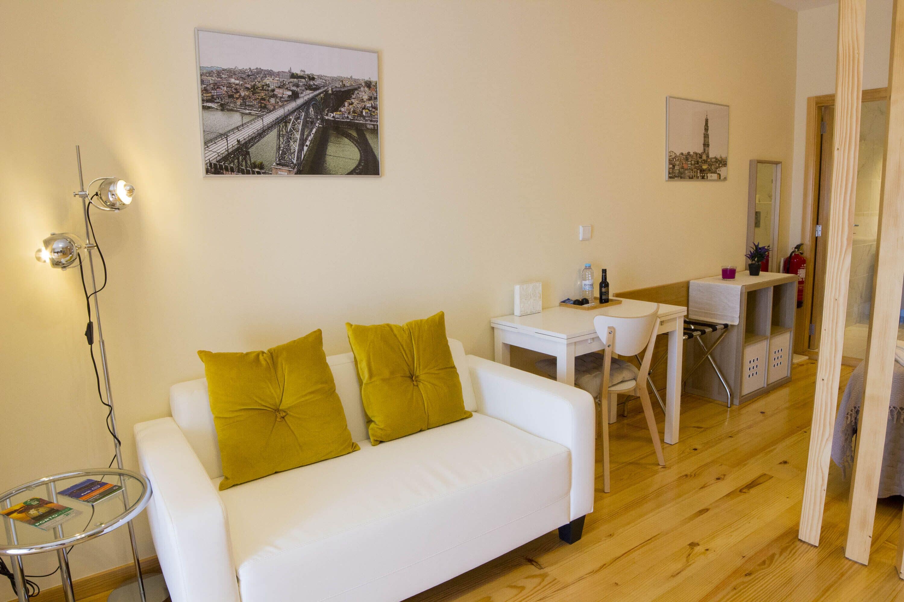 A modern living room with a white sofa, a glass coffee table, and a desk with a chair. There are two yellow cushions on the sofa.