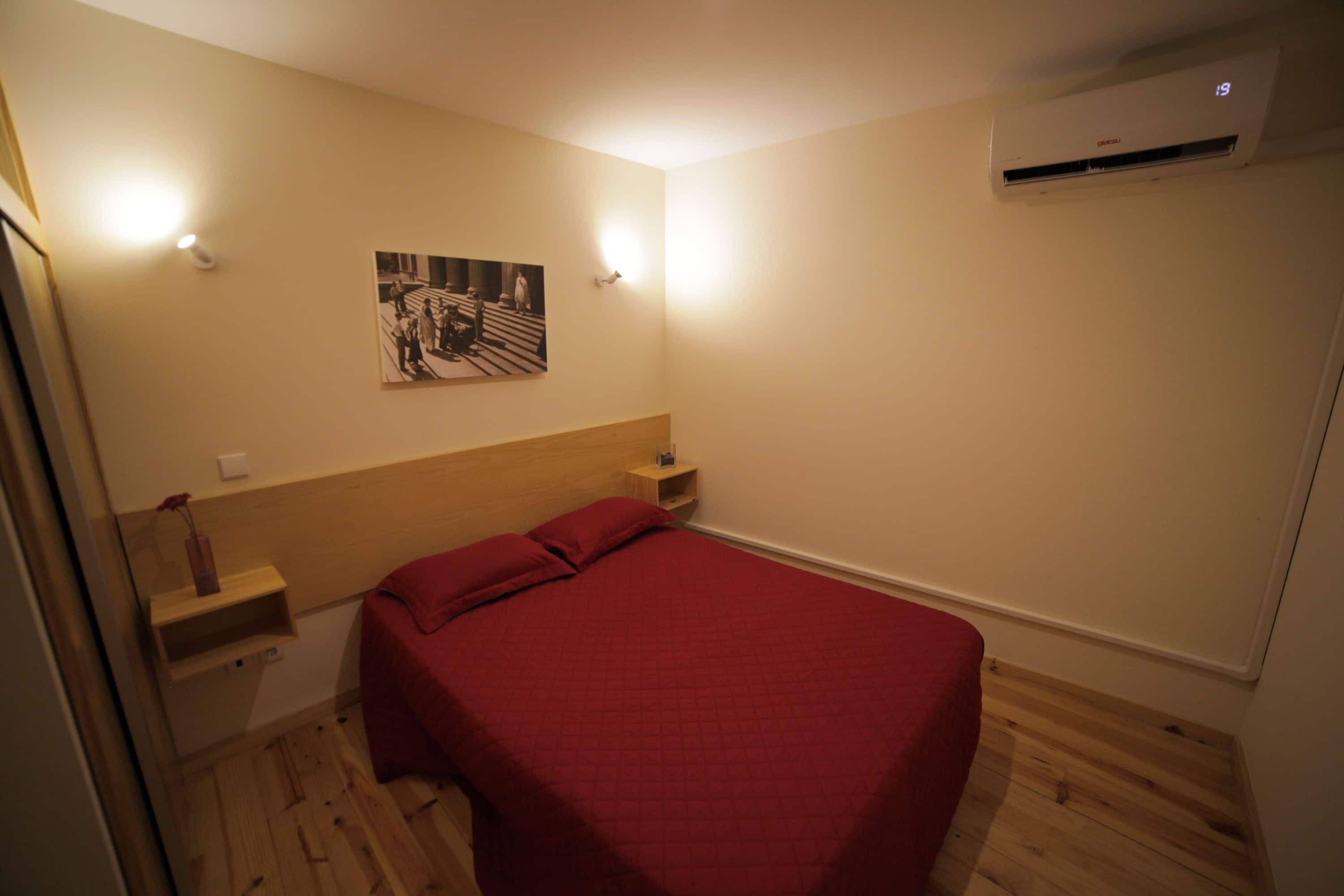 A bedroom with a wooden headboard, a red bedspread, a wall-mounted air conditioner, and a small shelf with a vase.