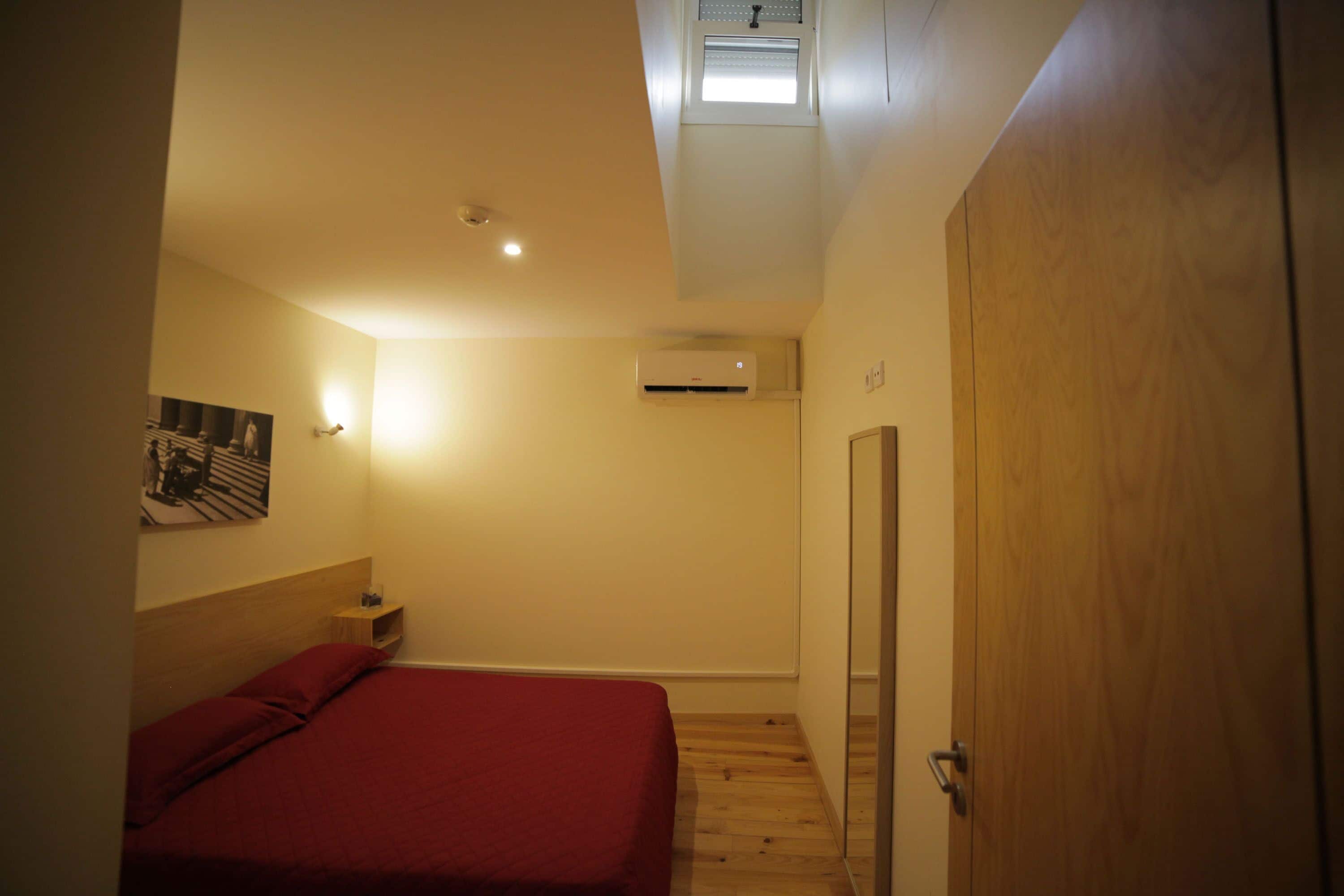 Un dormitorio con cama, mesita de noche, aire acondicionado, puerta de madera y tragaluz.