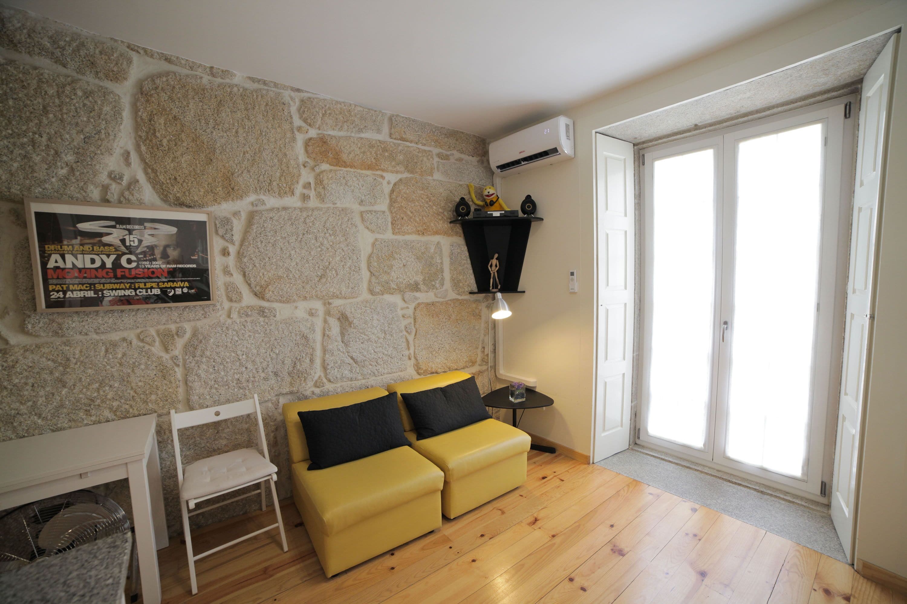 Una habitación con pared de piedra, un sofá amarillo, una pequeña silla blanca, una mesa con una lámpara y un cartel en la pared.