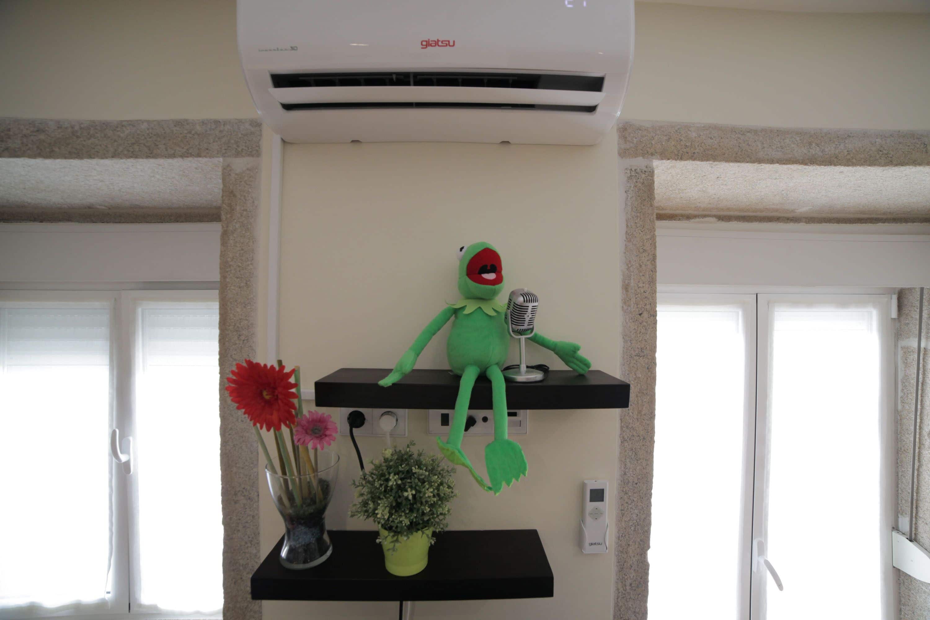 A room with a green plush toy on a shelf, a vase with flowers, and an air conditioning unit.