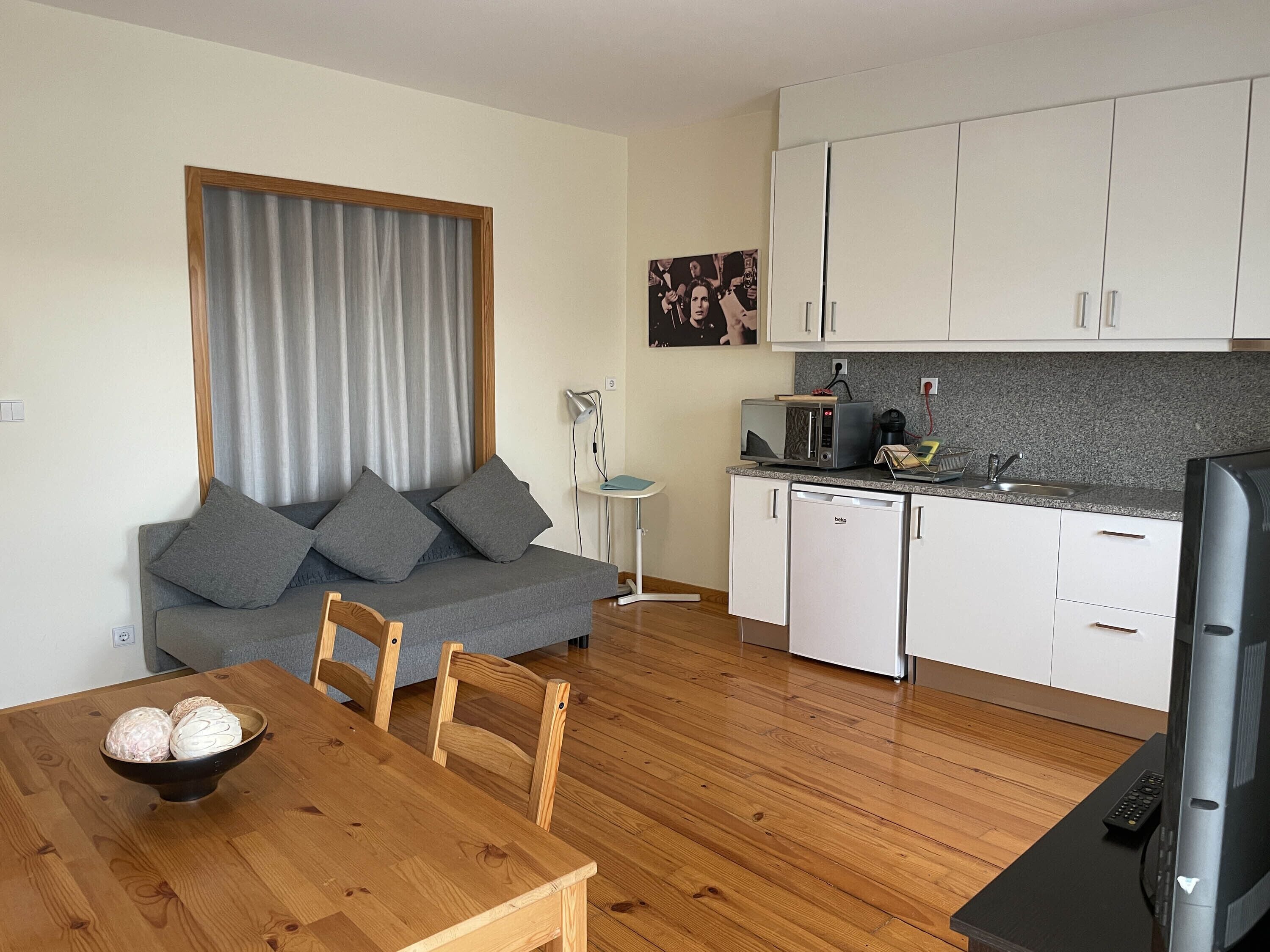 Un espacio habitable compacto con kitchenette, una zona de comedor con mesa y sillas de madera, un sofá gris con cojines y un televisor.