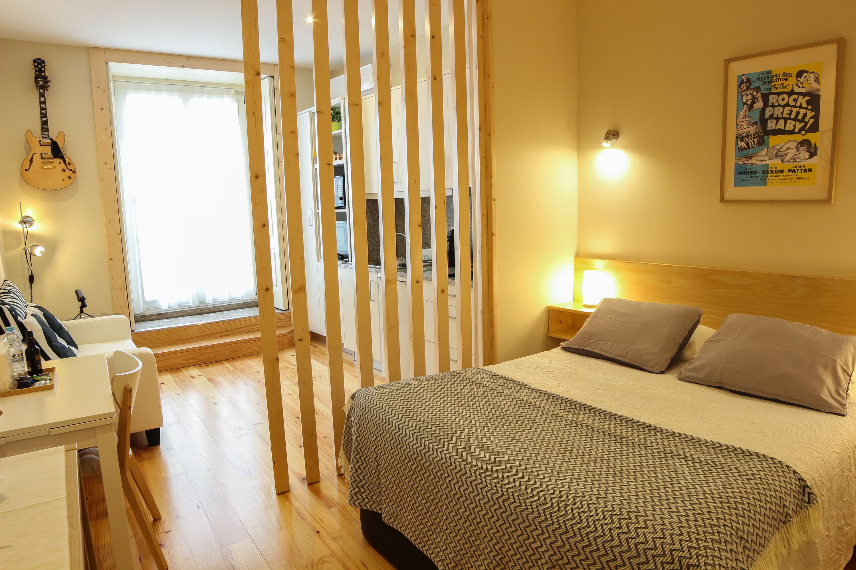A bedroom with a bed, pillows, a guitar, a desk, and a window.