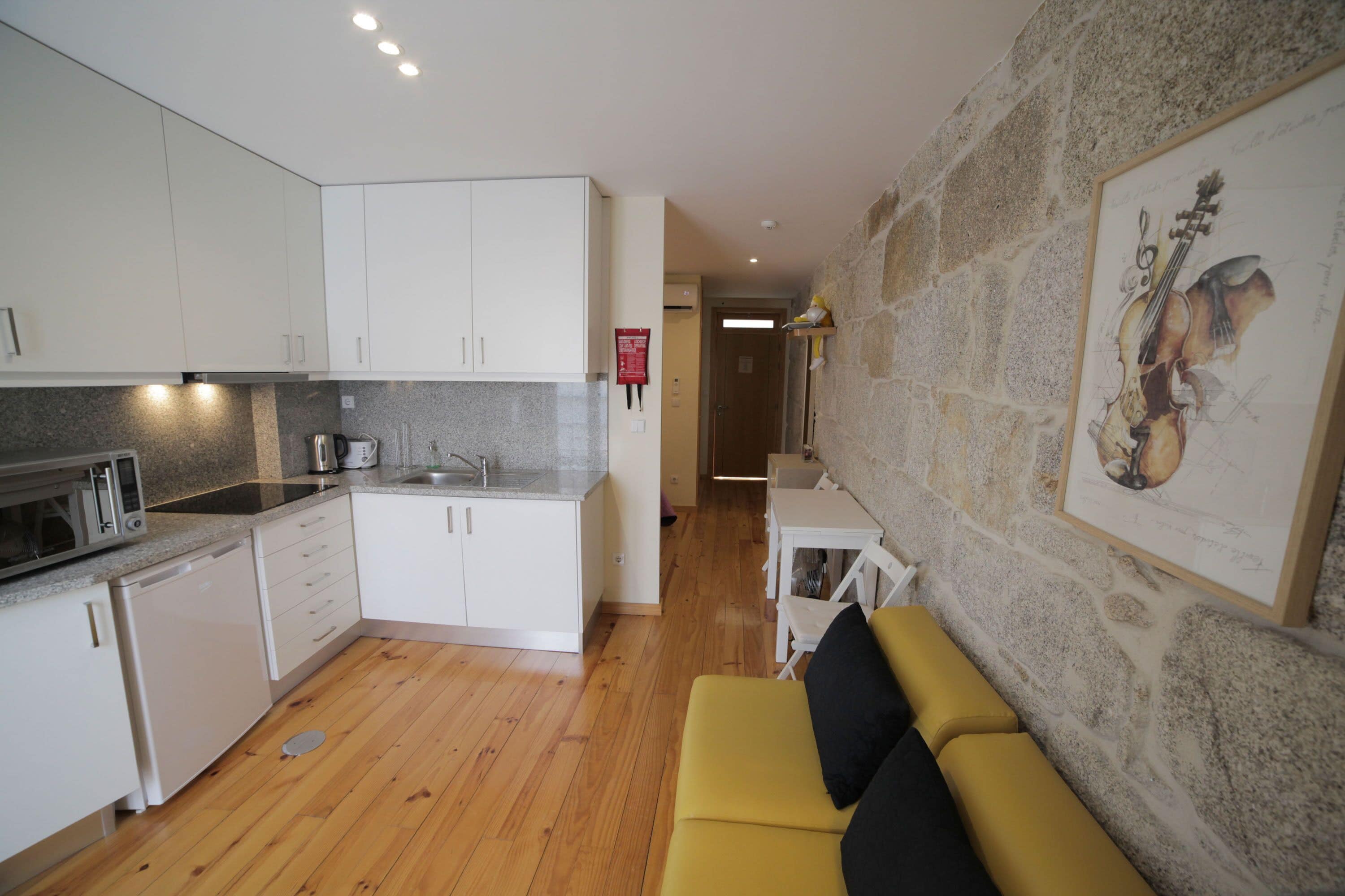 Un espacio habitable compacto con cocina, un sofá amarillo y una pared de piedra decorada con una obra de arte enmarcada.