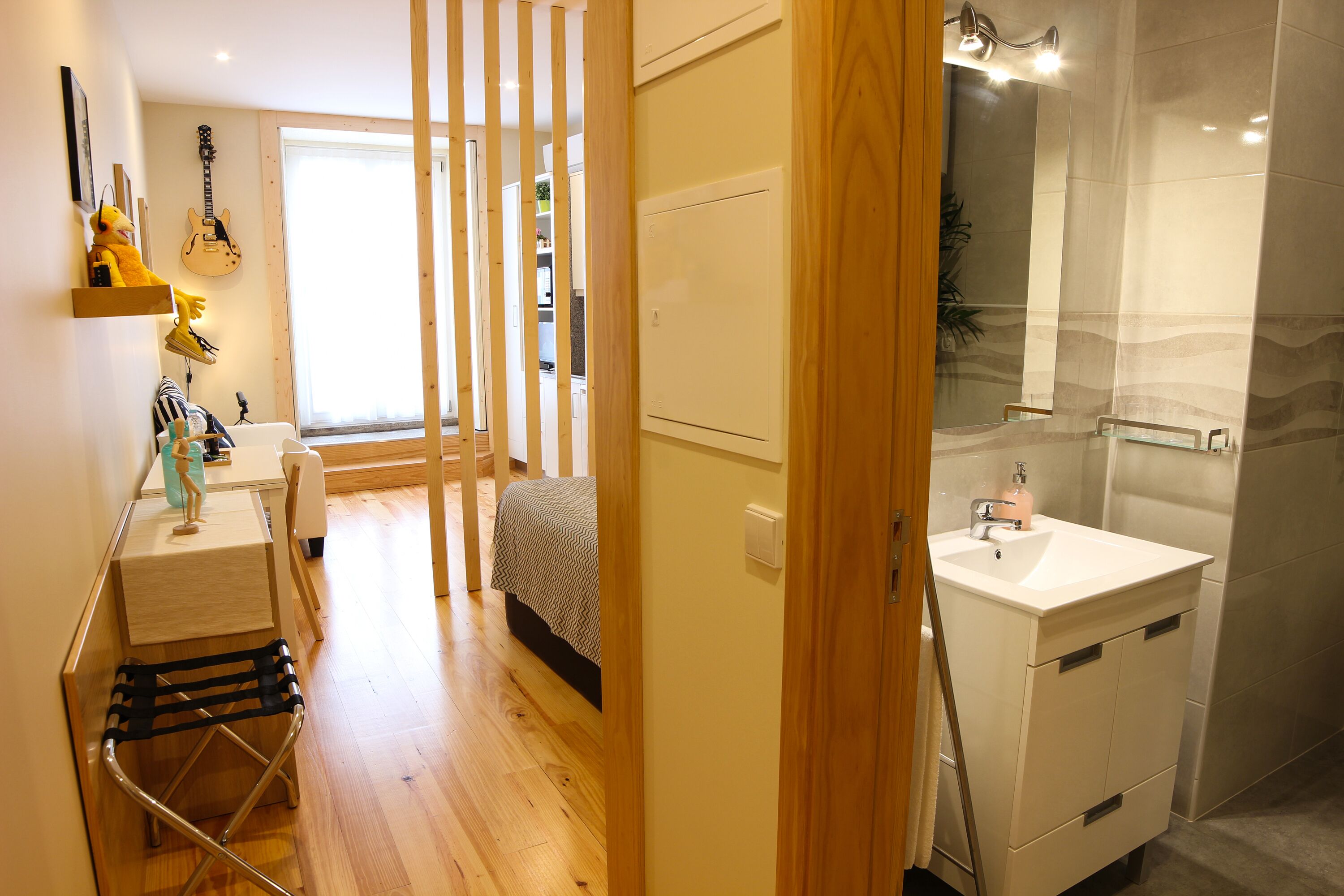 Une chambre à coucher de taille réduite, comprenant un lit, une table de chevet et un petit bureau avec une chaise. Une salle de bain avec un lavabo et un miroir est visible à travers une porte ouverte.