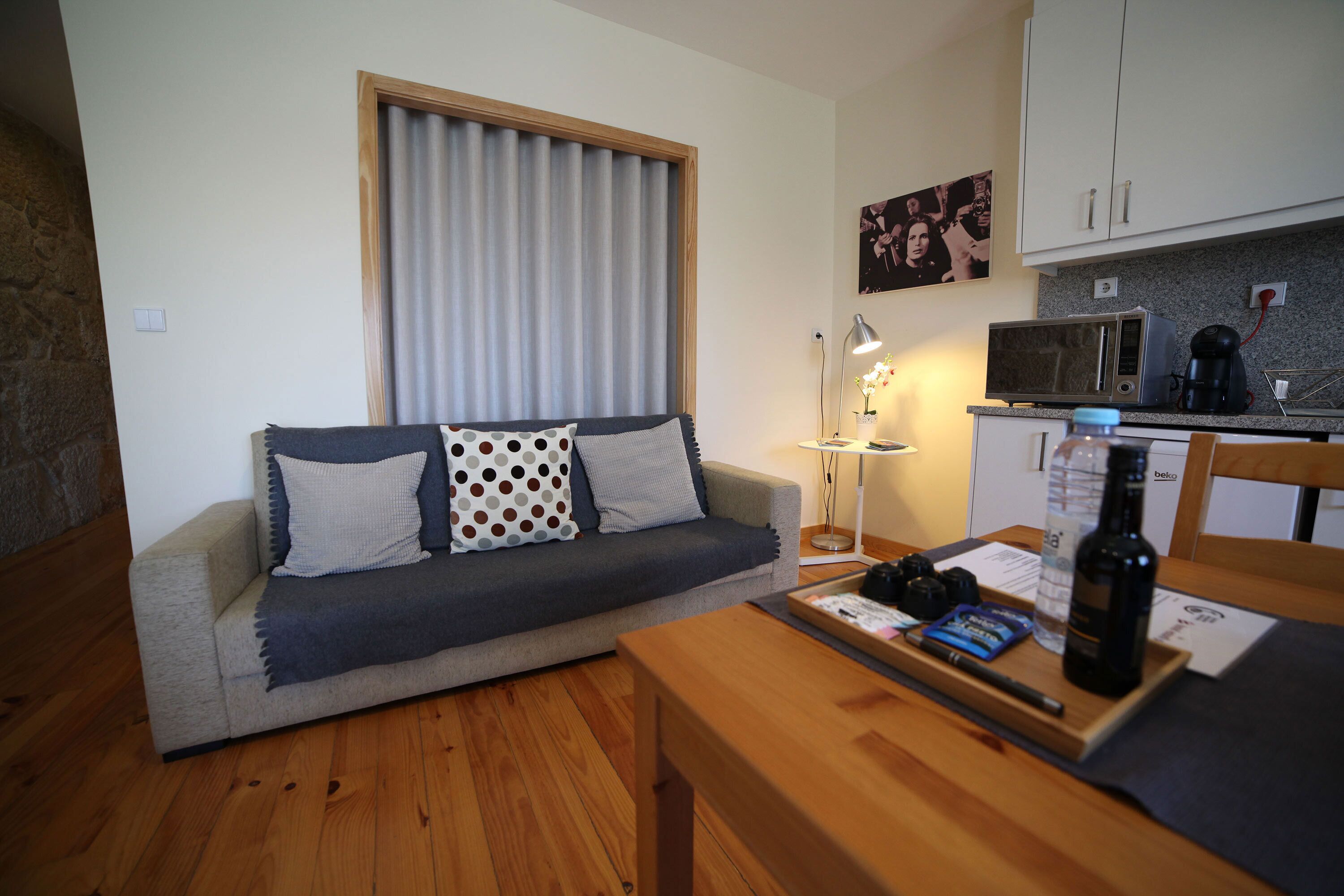 A compact living space with a sofa, a wooden table, a kitchen area with a microwave, and a window with curtains.